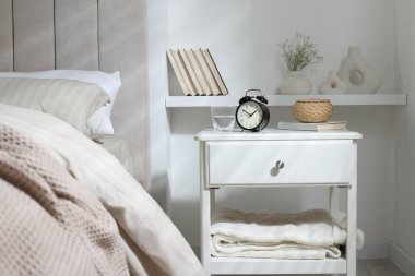 Alarm saati, bir bardak su, hasır kutu ve yatağın yanında kitap olan komodin.