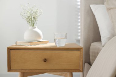 Komodinin yanında vazoda çiçekler, yatağın yanında bir bardak su ve kitap, yakın plan.