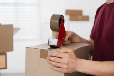 Karton kutuyu kapalı bir yere kapatmak için yapıştırıcılı bant kullanan bir adam.