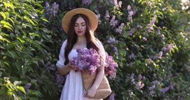 Açık havada çalıların yanında poz veren bir çanta dolusu leylak çiçeğiyle gülümseyen kadın.