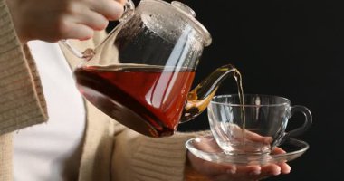 Kadın çaydanlıktan fincana bitki çayı dolduruyor. Siyah arka planda, yakın plan.