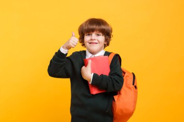 Sırt çantası ve kitabıyla turuncu arka planda baş parmağını gösteren sevimli bir okul çocuğu.