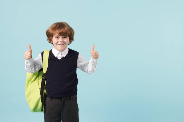 Sırt çantalı yakışıklı okul çocuğu açık mavi arkaplanda baş parmak gösteriyor. Mesaj için yer var.