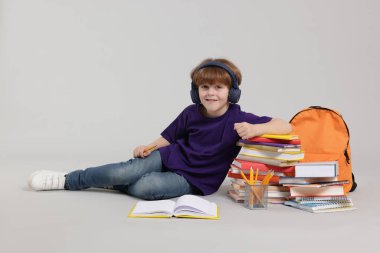 Sırt çantası, kitapları ve açık gri arka planda farklı kırtasiyeleri olan kulaklıklı tatlı bir okul çocuğu.