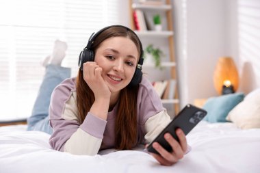 Kulaklıklı genç kız evde yatakta akıllı telefon kullanıyor.