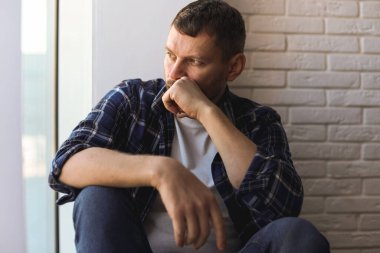 Evdeki pencerenin yanında oturan depresif bir adam.