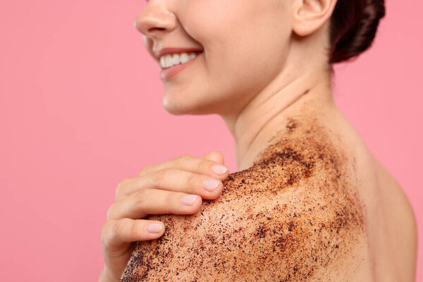Улыбающаяся женщина прикладывает скраб для тела к плечу на розовом фоне, крупным планом