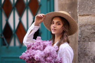 Şapkalı güzel bir kadın ve bina yakınlarında bir demet leylak çiçeği.