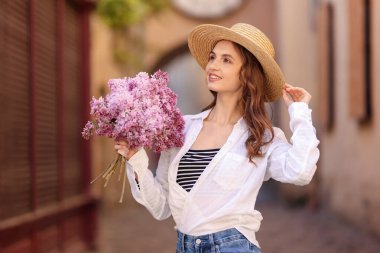 Şapkalı gülümseyen kadın, şehir caddesinde bir demet leylak çiçeğiyle.