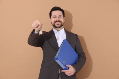 Emlakçının elinde ev şeklindeki anahtarlık ve bej arka planda klasör var.