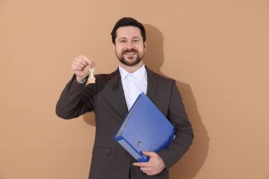 Emlakçının elinde ev şeklindeki anahtarlık ve bej arka planda klasör var.