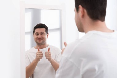 Kapalı alanda aynanın yanında baş parmağını gösteren yakışıklı adam.