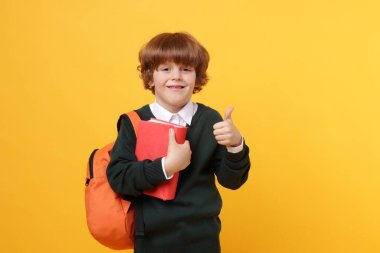 Sırt çantası ve kitabıyla turuncu arka planda baş parmağını gösteren sevimli bir okul çocuğu.