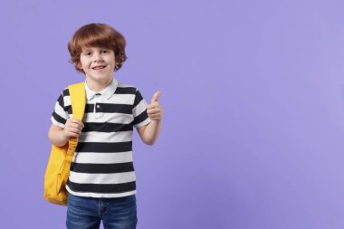 Sırt çantalı yakışıklı okul çocuğu menekşe arkaplanda baş parmak gösteriyor, mesaj için yer var.