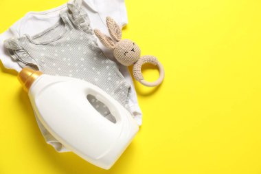 Bir şişe yumuşatıcı, sarı arka planda oyuncak ve bebek kıyafetleri, düz yatak. Metin için boşluk