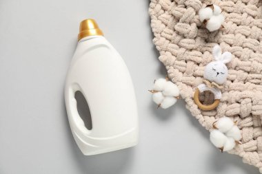 Bir şişe yumuşatıcı, oyuncak, bebek battaniyesi ve açık gri arka planda pamuk çiçekleri.