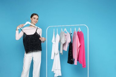 Gülümseyen moda tasarımcısı ve açık mavi arka planda giysileri olan askılar.