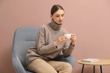 Güzel bir kadın, bir bardak içkiyle bej duvarın yanındaki sallanan sandalyede dinleniyor.