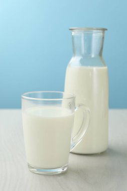 Bardakta taze süt ve açık mavi arka planda beyaz ahşap masada sürahi.