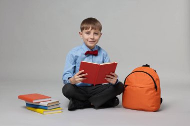 Sırt çantası açık gri arka planda kitap okuyan sevimli bir okul çocuğu.
