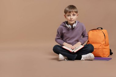 Kulaklıklı, sırt çantalı, kahverengi arka planda kitap okuyan tatlı bir okul çocuğu.