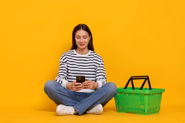 Sarı arka planda akıllı telefonu ve sepeti olan gülümseyen bir kadın. İnternetten alışveriş