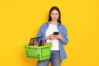Akıllı telefonu ve sarı arka planda ürünlerle dolu sepeti olan gülümseyen bir kadın. İnternetten alışveriş