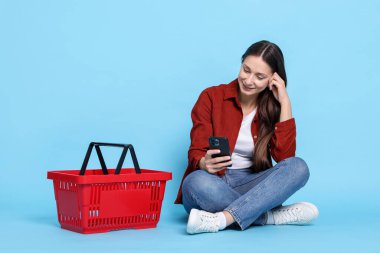 Açık mavi arka planda akıllı telefonu ve sepeti olan gülümseyen bir kadın. İnternetten alışveriş