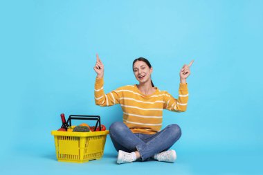 Gülümseyen kadın, açık mavi arka planda yukarı bakan ürünlerle dolu alışveriş sepetiyle.