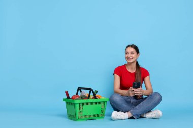 Ürünlerle dolu alışveriş sepeti ve açık mavi arka planda akıllı telefonu olan gülümseyen kadın. Metin için boşluk