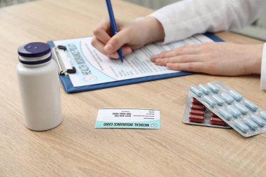 Doktor evrakları hazırlıyor, sağlık sigortası kartı ve haplar ahşap masada, yakın plan.