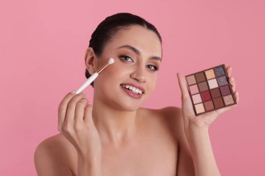 Göz farı paleti ve fırçasıyla gülümseyen kadın pembe arka plana makyaj yapıyor.