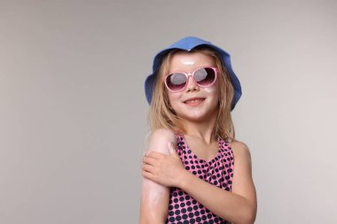 Gülümseyen küçük bir kız, gri arka planda omzuna güneş kremi sürüyor. Metin için boşluk