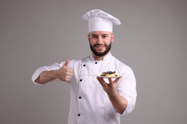 Üniformalı mutlu pastacı elinde yaban mersinli lezzetli bir tart ve açık gri arka planda baş parmak gösteriyor.
