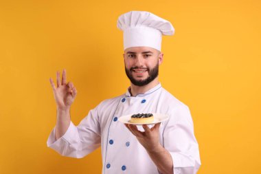 Üniformalı mutlu pastacı elinde yaban mersinli turta ve turuncu arka planda güzel bir jest gösteriyor.