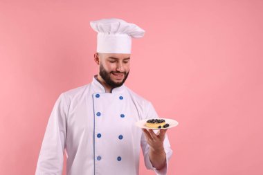 Üniformalı mutlu pastacı pembe arka planda yaban mersinli tart tutuyor.