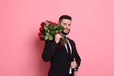 Kırmızı gül buketi olan mutlu bir adam pembe arka planda baş parmağını gösteriyor.