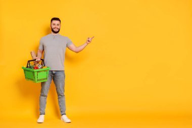 Elinde alışveriş sepeti dolusu mal olan adam turuncu arka plandaki bir şeyi işaret ediyor. Mesaj için yer var.