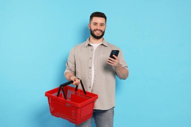 Boş alışveriş sepeti ve açık mavi arka planda akıllı telefonu olan bir adam.