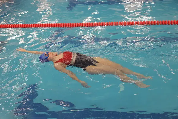 Kapalı havuzda yüzen şapkalı ve gözlüklü bir kadın.