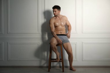 Beyaz duvarın yanındaki sandalyede açık gri iç çamaşırlı genç adam.