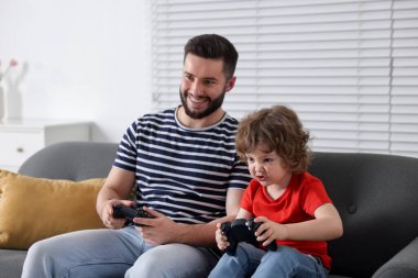 Mutlu baba ve oğlu evdeki kanepede oyun kumandalarıyla oynuyorlar.