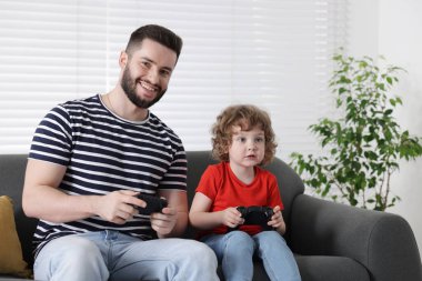 Mutlu baba ve oğlu evdeki kanepede oyun kumandalarıyla oynuyorlar.