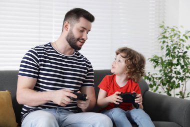 Mutlu baba ve oğlu evdeki kanepede oyun kumandalarıyla oynuyorlar.