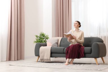 Kadın pencerenin yanındaki kanepede kitap okuyor ve evinde şık perdeler var. Metin için boşluk