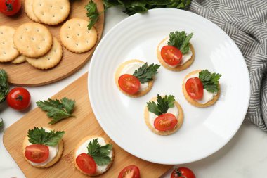 Krem peynirli krakerler, domatesler ve maydanozlar beyaz masada.