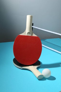 Masa tenisi raketleri, top ve ağ açık mavi masada.