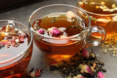 Cam fincanlarda aromatik bitki çayı, gri masada kuru yapraklar ve çiçekler, yakın plan.