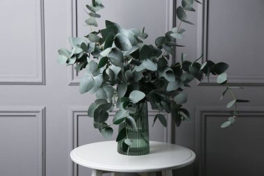 Gri duvarın yanındaki masada vazoda okaliptüs dalları var.