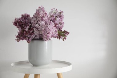 Beyaz duvarın yanındaki masada vazodaki güzel leylak çiçekleri, metin için yer var.